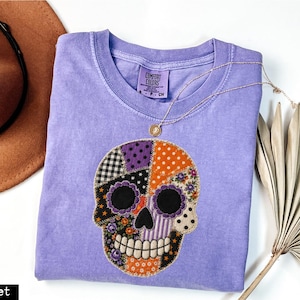 Puede incluir: Una camiseta violeta de manga corta con un gráfico central de calavera estilo patchwork. La calavera está compuesta por varios segmentos de tela estampada en naranja, morado, negro y blanco. La camiseta tiene una etiqueta Comfort Colors en el cuello.