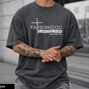 Puede incluir: Camiseta de gran tamaño color pimienta con el texto "FATHERHOOD IS KINGDOM WORK" y un gráfico de cruz. La camiseta también incluye el texto "PROVERBIOS 22:6". La camiseta es de color gris oscuro y tiene un aspecto vintage y desgastado.