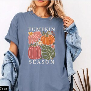 May include: A blue jean colored t-shirt featuring a graphic print with the text PUMPKIN SEASON. The design includes illustrations of two pumpkins, a large pink flower, a green monstera leaf, and small floral accents in orange and white.