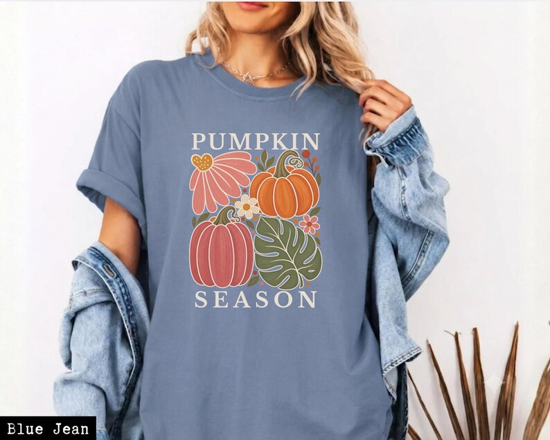 May include: A blue jean colored t-shirt featuring a graphic print with the text PUMPKIN SEASON. The design includes illustrations of two pumpkins, a large pink flower, a green monstera leaf, and small floral accents in orange and white.