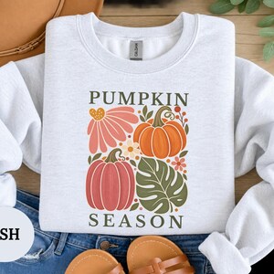 May include: An ash gray crewneck sweatshirt featuring a graphic print with the text PUMPKIN SEASON. The design includes a large orange pumpkin, a smaller pink pumpkin, a pink flower, a green monstera leaf, and small floral accents in autumn colors.