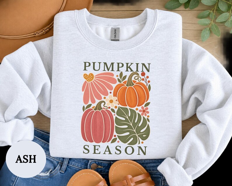 May include: An ash gray crewneck sweatshirt featuring a graphic print with the text PUMPKIN SEASON. The design includes a large orange pumpkin, a smaller pink pumpkin, a pink flower, a green monstera leaf, and small floral accents in autumn colors.