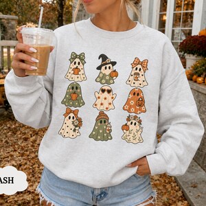 Puede incluir: Una sudadera gris ceniza de cuello redondo con un estampado gráfico de nueve fantasmas distintos. Cada fantasma está decorado con diferentes patrones y accesorios de temática otoñal, incluyendo calabazas, flores, lazos y sombreros.