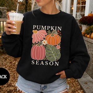 May include: A black long-sleeve sweatshirt featuring a graphic print with the text PUMPKIN SEASON. The design includes illustrations of two pumpkins, a large pink flower, a green monstera leaf, and small white flowers in a grid layout.