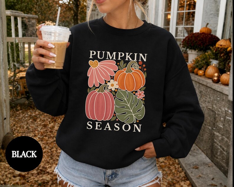 May include: A black long-sleeve sweatshirt featuring a graphic print with the text PUMPKIN SEASON. The design includes illustrations of two pumpkins, a large pink flower, a green monstera leaf, and small white flowers in a grid layout.