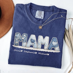 Puede incluir: Camiseta azul marino con la palabra "MAMA" en un diseño de patchwork con patrones florales y de corazón azules. Los nombres Olivia, Stephanie y Michael están impresos debajo. También se ve un collar dorado y un sombrero marrón.