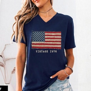 Puede incluir: Camiseta azul marino con cuello en V con un diseño de bandera estadounidense desgastada. El diseño de la bandera incluye estrellas y rayas, con el texto "VINTAGE 1976" debajo. Una prenda casual y patriótica.