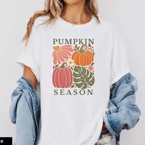 May include: A white short-sleeve t-shirt featuring a graphic print with the text PUMPKIN SEASON. The design includes illustrations of orange and pink pumpkins, a large green monstera leaf, and small flowers in shades of pink and white.