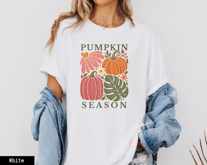 May include: A white short-sleeve t-shirt featuring a graphic print with the text PUMPKIN SEASON. The design includes illustrations of orange and pink pumpkins, a large green monstera leaf, and small flowers in shades of pink and white.