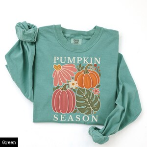 May include: A light green long-sleeve crewneck sweatshirt featuring a graphic print with the text PUMPKIN SEASON. The design includes two pumpkins, a large pink flower, a small white flower, and a green monstera leaf in a central arrangement.