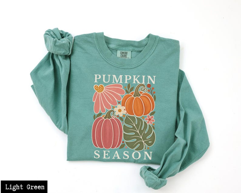 May include: A light green long-sleeve crewneck sweatshirt featuring a graphic print with the text PUMPKIN SEASON. The design includes two pumpkins, a large pink flower, a small white flower, and a green monstera leaf in a central arrangement.