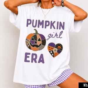 Puede incluir: Una camiseta blanca de manga corta con el texto morado PUMPKIN GIRL ERA. El gráfico incluye una calabaza estilo patchwork y una forma de corazón, ambos decorados con patrones naranjas, morados y negros que incluyen fantasmas y murciélagos.