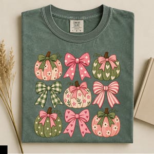 Puede incluir: Una camiseta verde musgo con un estampado gráfico de tres calabazas y tres lazos dispuestos en cuadrícula. Las calabazas están decoradas con motivos florales, corazones y lunares en tonos rosa y verde, combinados con varios lazos.