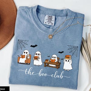 Puede incluir: Una camiseta color azul vaquero con un gráfico de cuatro fantasmas blancos leyendo libros y bebiendo café. El texto "the boo club" está impreso debajo en letra cursiva blanca. La camiseta tiene cuello redondo y una etiqueta Comfort Colors.