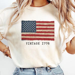 Puede incluir: Camiseta de color marfil con un diseño de bandera estadounidense desgastada con rayas rojas y blancas y un campo de estrellas azul. El texto "VINTAGE 1776" está impreso debajo. La camiseta tiene mangas cortas.