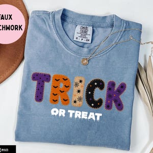 Puede incluir: Una camiseta color azul jean con un diseño de falso patchwork que muestra el texto TRICK en letras coloridas con patrones y el texto blanco OR TREAT debajo. La camiseta tiene cuello redondo y una etiqueta de la marca Comfort Colors.