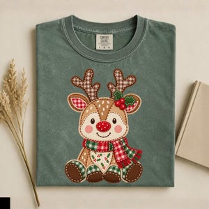 Puede incluir: Una camiseta de cuello redondo color verde musgo con un gráfico central de un reno de dibujos animados con pelaje marrón y blanco, nariz roja y bufanda a cuadros. El reno tiene astas de cuadros marrones y está decorado con bayas de acebo rojas.