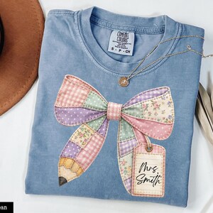 Puede incluir: Una camiseta azul vaquera Comfort Colors con un diseño de lazo y lápiz de patchwork. El lazo incorpora patrones rosas, morados y florales. Se adjunta una etiqueta con "Mrs. Smith". También se ven un collar dorado y un sombrero marrón.