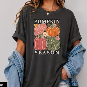 May include: A charcoal gray short-sleeve t-shirt featuring a graphic print with the text PUMPKIN SEASON. The design includes two pumpkins, a large pink flower, a green monstera leaf, and small white flowers arranged in a square composition.