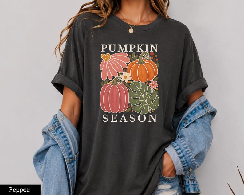 May include: A charcoal gray short-sleeve t-shirt featuring a graphic print with the text PUMPKIN SEASON. The design includes two pumpkins, a large pink flower, a green monstera leaf, and small white flowers arranged in a square composition.