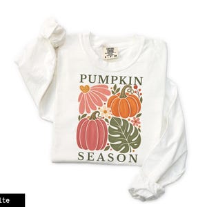 May include: A white long-sleeve t-shirt featuring a graphic print with the text PUMPKIN SEASON in dark green letters. The design includes two orange and pink pumpkins, a large pink flower, a green monstera leaf, and small floral accents.