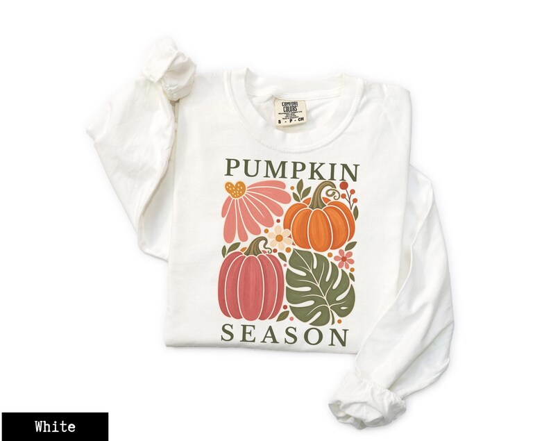 May include: A white long-sleeve t-shirt featuring a graphic print with the text PUMPKIN SEASON in dark green letters. The design includes two orange and pink pumpkins, a large pink flower, a green monstera leaf, and small floral accents.