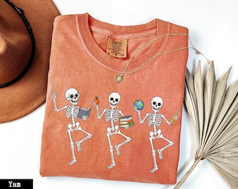 May include: A burnt orange t-shirt featuring a graphic of three white skeletons in various poses. The first skeleton holds a pointer and a book, the second holds an apple and a stack of books, and the third holds a globe and a pencil.
