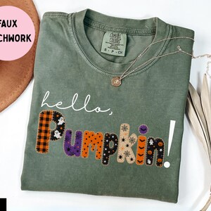 Puede incluir: Una camiseta de manga corta color verde musgo con el texto blanco hello, y la palabra PUMPKIN! en un diseño de imitación patchwork. Cada letra de la palabra PUMPKIN! muestra un patrón diferente con temática de Halloween, incluyendo fantasmas, calabazas y arañas.