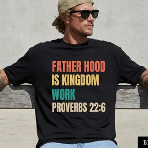 Puede incluir: Camiseta negra con el texto "FATHER HOOD IS KINGDOM WORK PROVERBS 22:6" en naranja, amarillo, turquesa y blanco roto. La camiseta es de cuello redondo.