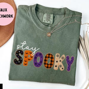 Puede incluir: Una camiseta de manga corta color verde musgo con el texto cursivo blanco stay sobre la palabra SPOOKY en letras de imitación patchwork. Cada letra tiene un patrón distinto que incluye murciélagos, cuadros, arañas y telarañas.