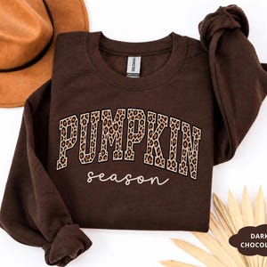 Puede incluir: Una sudadera de cuello redondo en color marrón chocolate oscuro con la palabra PUMPKIN en una fuente universitaria con estampado de leopardo, y la palabra season escrita debajo en letra cursiva blanca. La prenda tiene mangas largas y cuello acanalado.