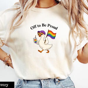 Puede incluir: Camiseta de color marfil con un dibujo de un ganso con gafas de sol y un sombrero morado, sosteniendo una bandera arcoíris y una bebida. El texto "Off to Be Proud" está encima del ganso. La camiseta es de material suave.