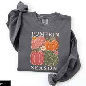 May include: A charcoal gray long-sleeve sweatshirt featuring a graphic print with the text PUMPKIN SEASON. The design includes two pumpkins in orange and muted red, a large pink flower, a green monstera leaf, and small white flowers.