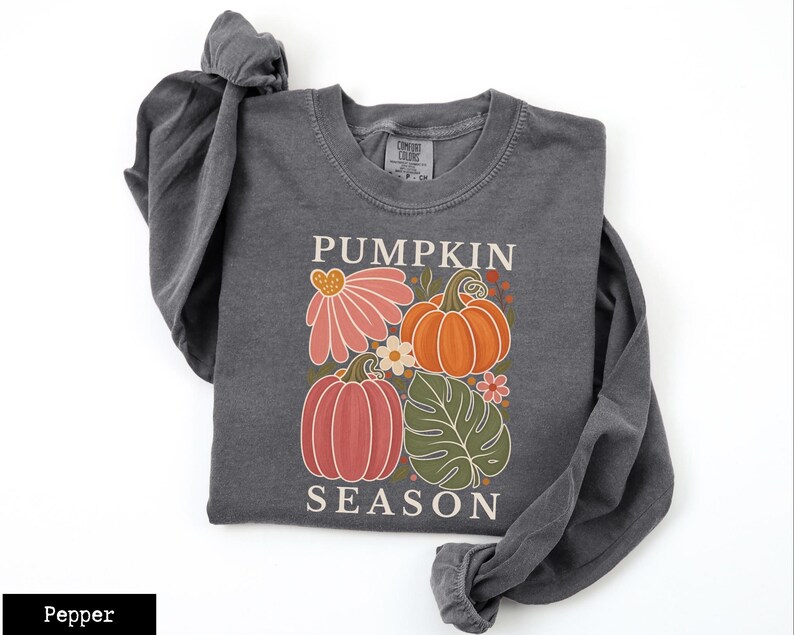 May include: A charcoal gray long-sleeve sweatshirt featuring a graphic print with the text PUMPKIN SEASON. The design includes two pumpkins in orange and muted red, a large pink flower, a green monstera leaf, and small white flowers.