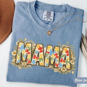 Puede incluir: Camiseta azul vaquero Comfort Colors con la palabra "MAMA" en piezas de rompecabezas coloridas, rodeada de detalles florales. Un collar dorado descansa sobre la camisa. La camiseta es talla S-P-CH.