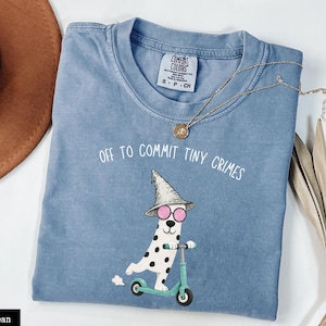 Puede incluir: Una camiseta de color azul vaquero con un gráfico de un dálmata con sombrero puntiagudo y gafas de sol rosas montado en un patinete verde claro. El texto sobre el gráfico dice OFF TO COMMIT TINY CRIMES en letras blancas.