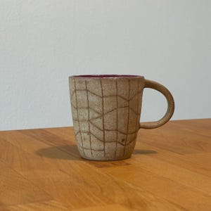 May include: A ceramic mug with a speckled beige exterior featuring a dark brown grid pattern of intersecting lines. The mug has a curved handle on the right side and a smooth, solid dark magenta interior finish that is visible at the top rim.