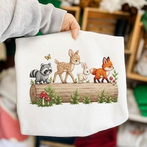Puede incluir: Sudadera blanca con un diseño de animales del bosque. Un mapache, un ciervo, un conejo y un zorro están representados en un tronco, con una mariposa, setas y follaje. La sudadera es sostenida por una persona.