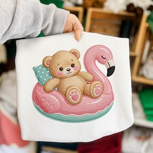 Puede incluir: Sudadera blanca con un diseño bordado de un oso de peluche relajándose en un flotador de flamenco rosa. El oso tiene mejillas rosadas y descansa sobre una almohada azul. El flotador de flamenco tiene una base turquesa.