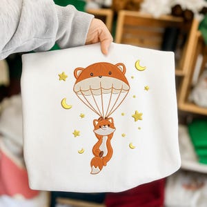 May include: White sweatshirt featuring an embroidered design of a fox suspended from a parachute shaped like a bear's head. The design includes gold stars and crescent moons. The fox is orange and white.