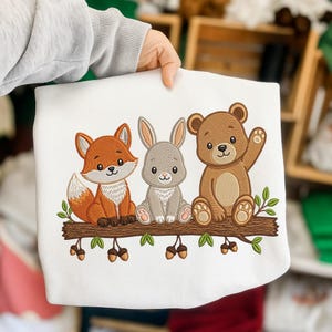 May include: White sweatshirt featuring an embroidered design of a fox, rabbit, and bear sitting on a branch with acorns. The fox is orange, the rabbit is gray, and the bear is brown. The design is detailed with green leaves.