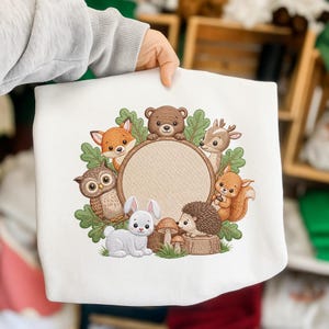 Könnte beinhalten: Weißes Sweatshirt mit einem kreisförmigen Design mit Waldtieren. Das Design zeigt einen Fuchs, einen Bären, ein Reh, ein Eichhörnchen, eine Eule, ein Kaninchen und einen Igel um einen leeren Kreis. Die Tiere sind in Braun-, Orange- und Weißtönen gehalten.