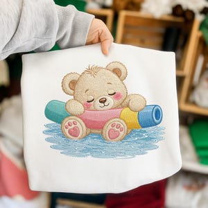 May include: White sweatshirt featuring an embroidered design of a sleeping teddy bear. The bear is light brown with pink accents, resting on a colorful, cylindrical object. The design is set against a blue, water-like background.