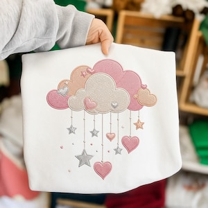 May include: White sweatshirt featuring an embroidered design of a cloud with pink, peach, and tan accents. The cloud has dangling stars and hearts in shades of pink and silver. The design is centered on the front of the sweatshirt.
