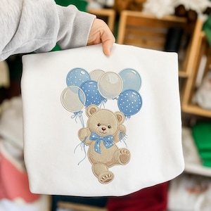May include: White sweatshirt featuring a cartoon teddy bear holding blue and white balloons. The bear has a blue bow tie. The balloons are in shades of blue and white. The design is an embroidered graphic.
