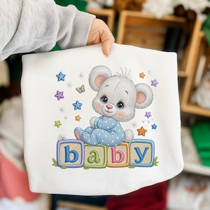 Puede incluir: Manta de bebé blanca con un oso de dibujos animados en pijama azul sentado sobre bloques que deletrean "baby". El diseño incluye estrellas, mariposas y detalles florales. Un diseño encantador y caprichoso para una guardería.