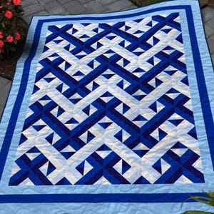 May include: A blue and white quilt with a geometric pattern. The quilt features a repeating design of interlocking shapes in shades of blue and white. The quilt has a light blue border and is displayed outdoors.