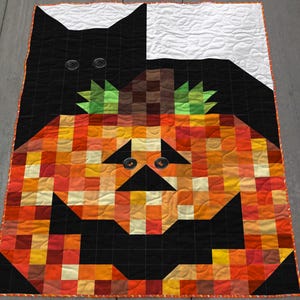 May include: A Halloween-themed quilt featuring a black cat and a jack-o'-lantern design. The quilt is made of squares in shades of orange, yellow, red, and black. The cat's eyes and the jack-o'-lantern's eyes are represented by buttons.