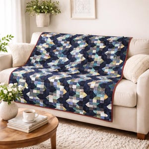 May include: A rectangular quilt featuring a geometric pattern of dark navy blue, light blue, teal, and cream-colored squares arranged in a repeating zigzag design. The quilt has a thin dark red border and is draped over a beige sofa.