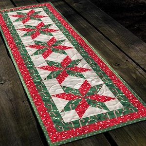 May include: A rectangular Christmas table runner with a repeating pattern of red and green star shapes on a white background. The runner is bordered with red fabric and candy cane accents, and measures approximately 30 cm wide and 122 cm long.